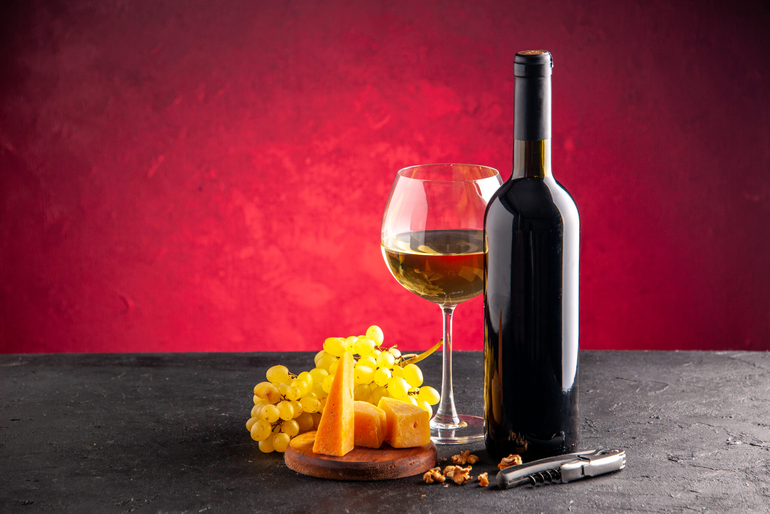 front view wine glass wine bottle yellow grapes cheese wood board wine opener dark table light red background scaled